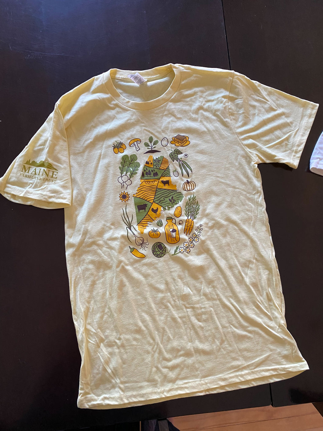 Light yellow t-shirt with illustrated yellow and green outline of Maine covered by rolling farmland, surrounded by yellow and green fruits, vegetables and other farm products. Olive green Maine Farmland Trust logo on the right sleeve.